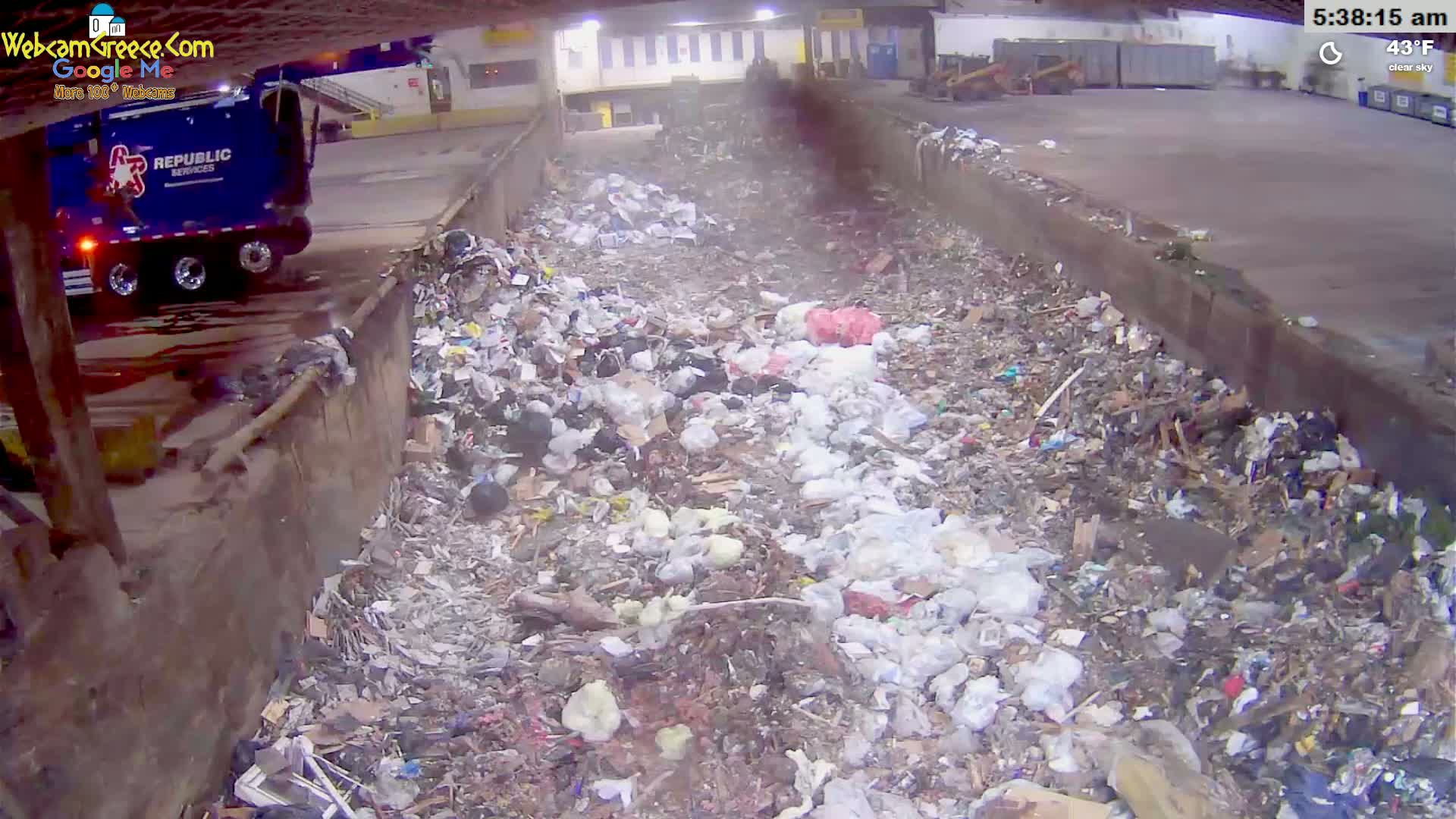 A garbage truck is parked near a large pile of trash in an indoor facility under clear skies and a temperature of 43°F.