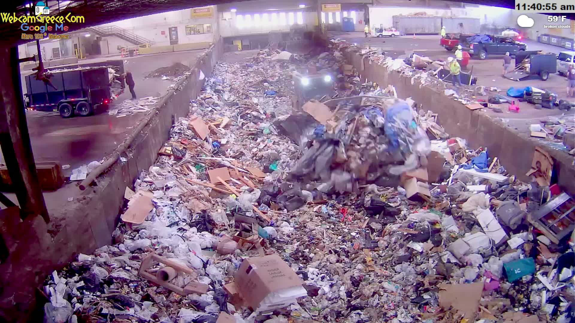 A large pile of trash is being compacted by machinery in an outdoor landfill under broken clouds and 59°F weather.