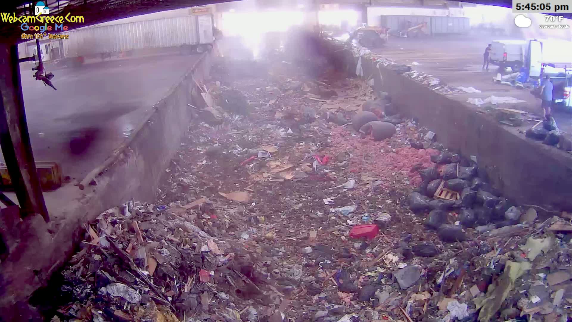 A large pile of trash and debris fills a concrete trough under an overcast sky, with several people visible in the background.
