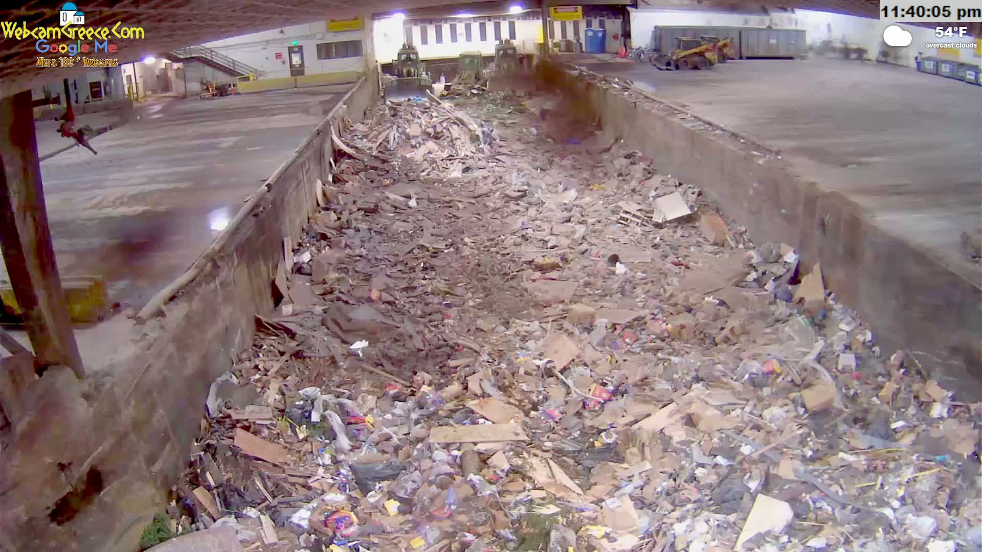 A large pile of refuse fills a long, concrete trough-like structure in an indoor facility under overcast skies.