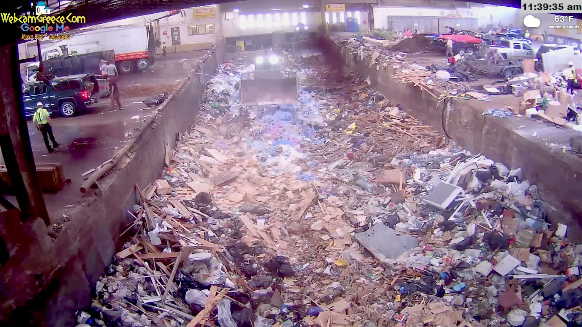 A large amount of refuse is piled high in an outdoor waste disposal area on an overcast day, with several people and vehicles visible nearby.