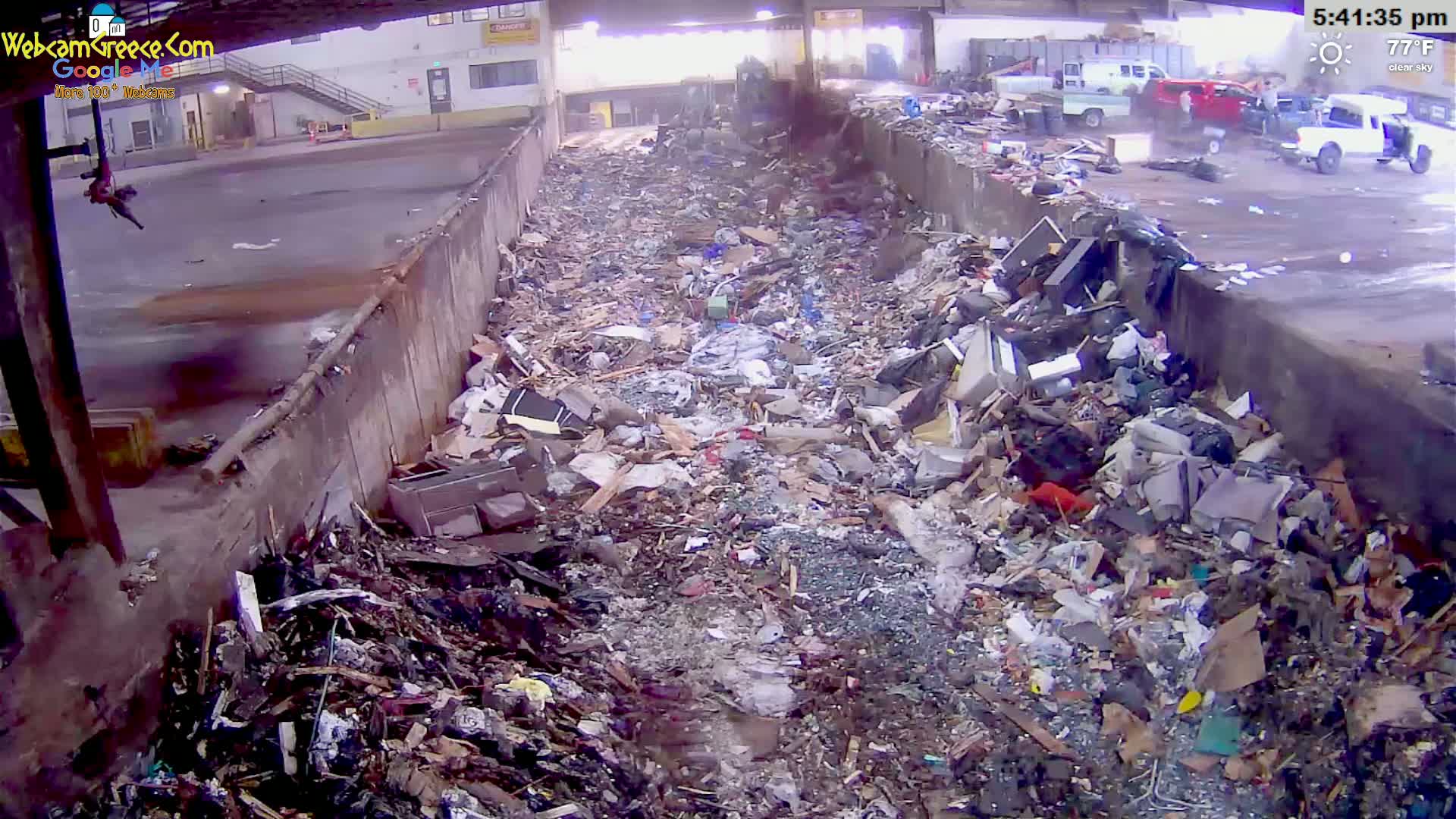 A large pile of discarded waste and debris fills a long, narrow concrete trough outdoors on a clear, 77°F day.