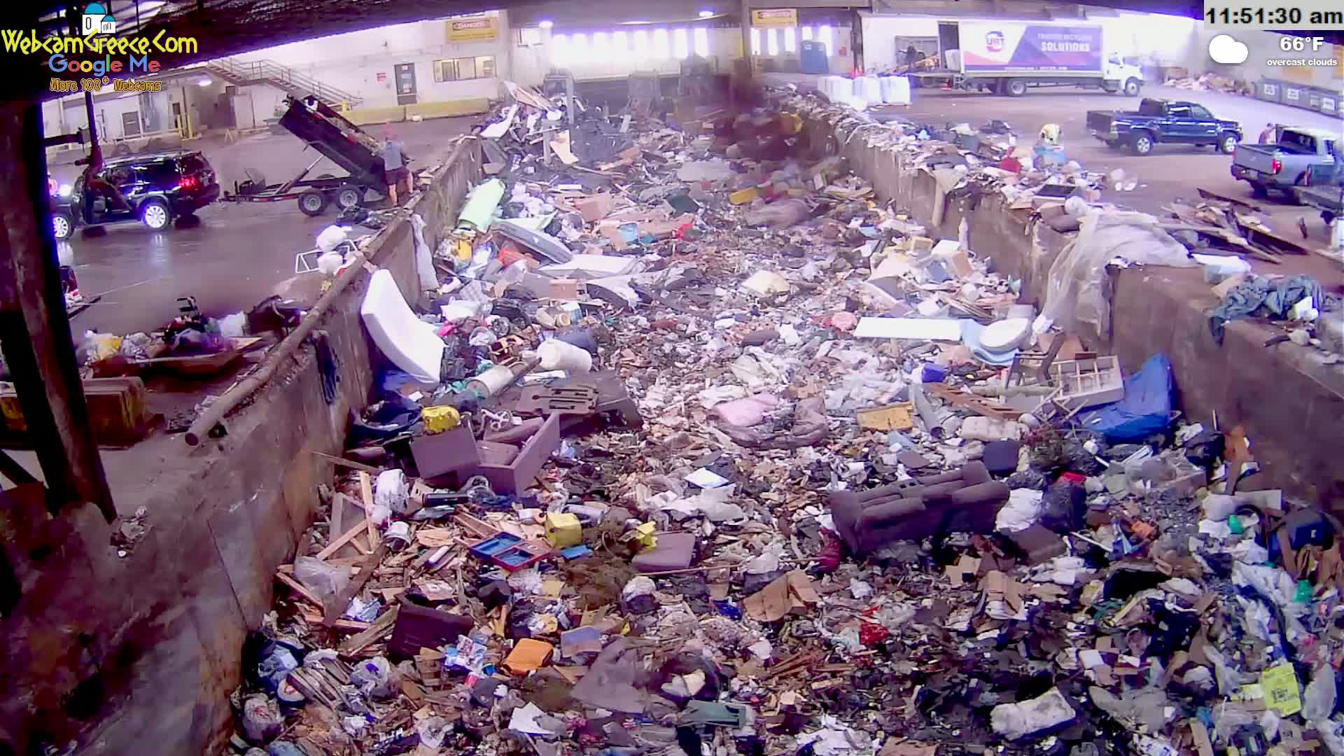 A large pile of refuse is seen in an indoor waste facility under overcast skies, with vehicles visible in the background.