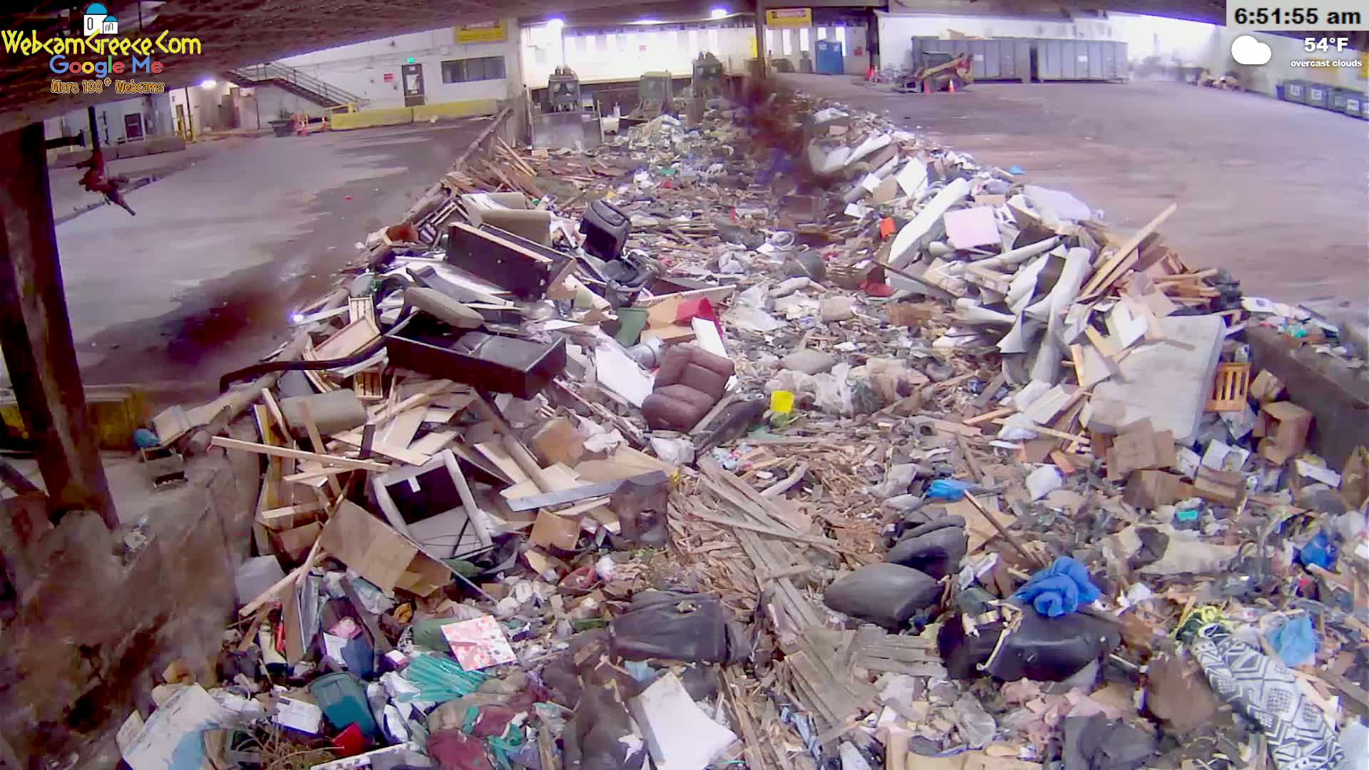 A large pile of discarded furniture and debris sits in an indoor waste facility under overcast skies.