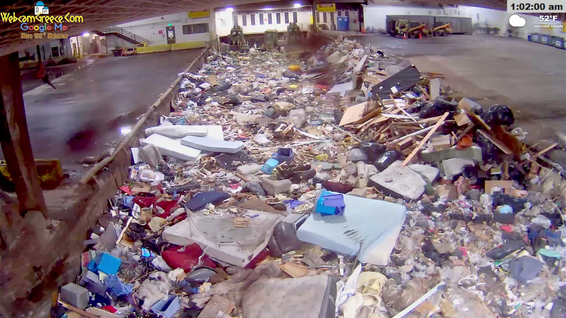 A large pile of discarded furniture, wood, and other refuse sits in an indoor waste facility under overcast skies.