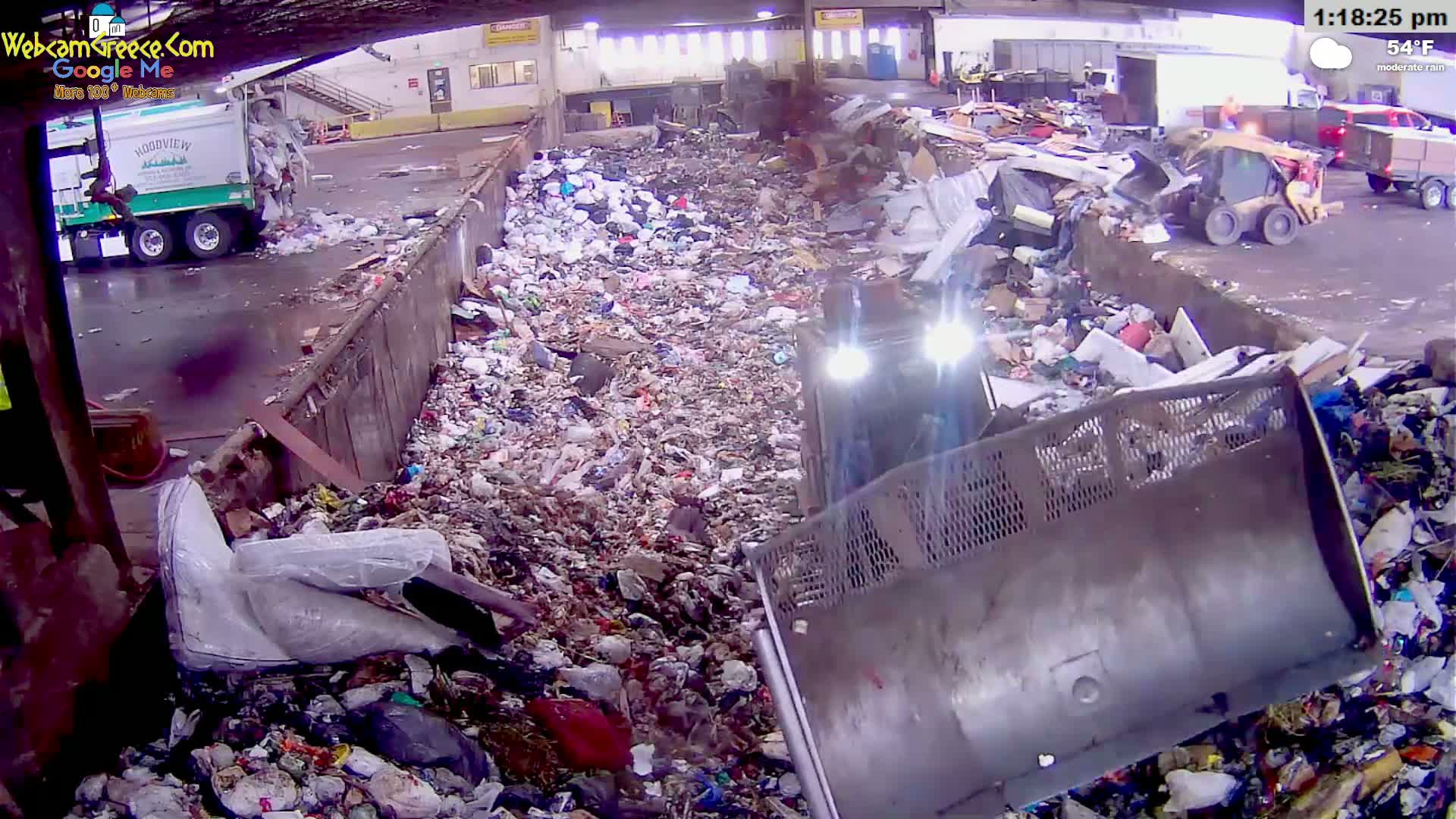 A large amount of garbage is being processed by machinery under moderate rain in a waste processing facility.