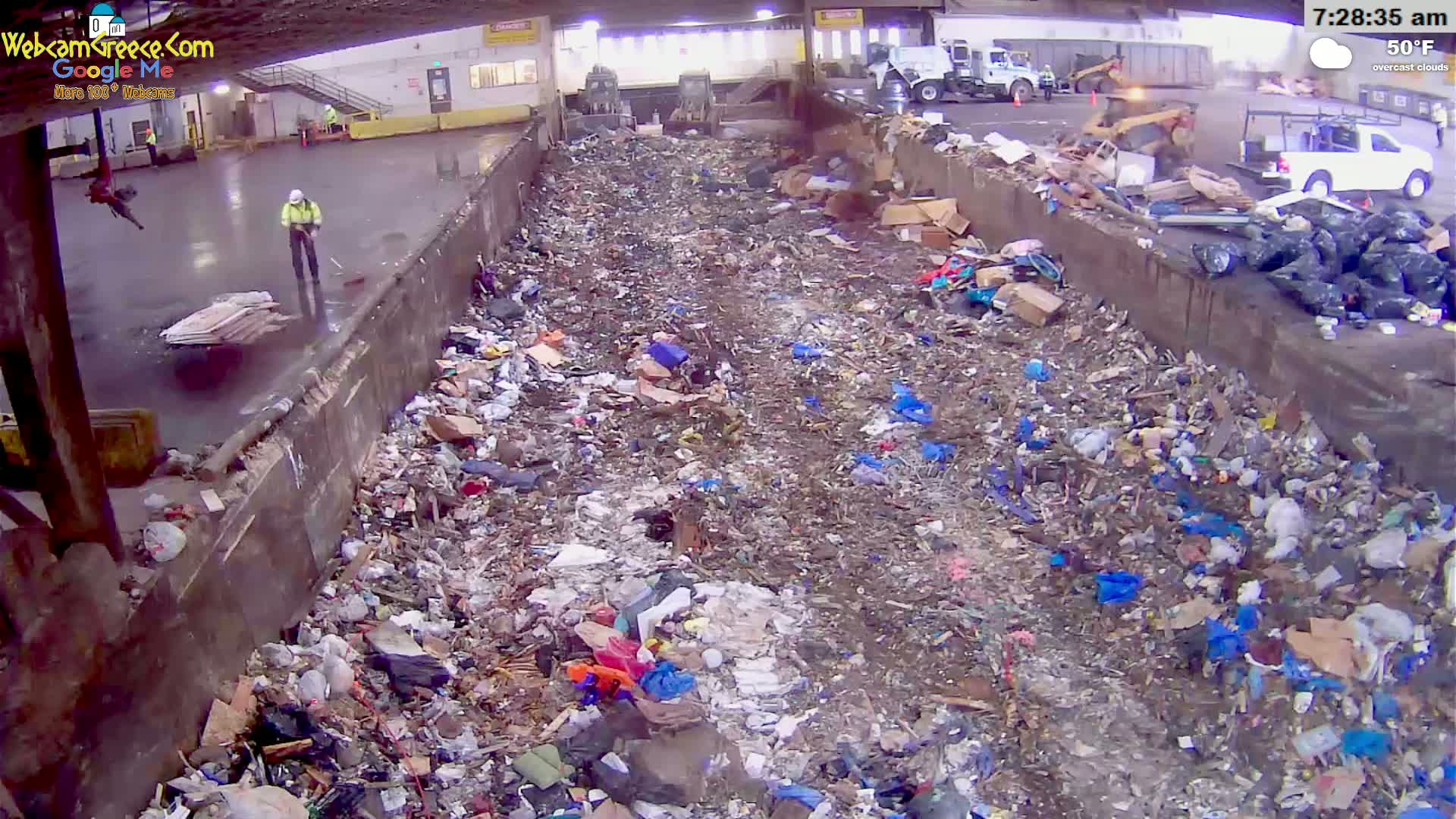 A worker sweeps a wet concrete floor next to a large pile of trash under overcast skies.