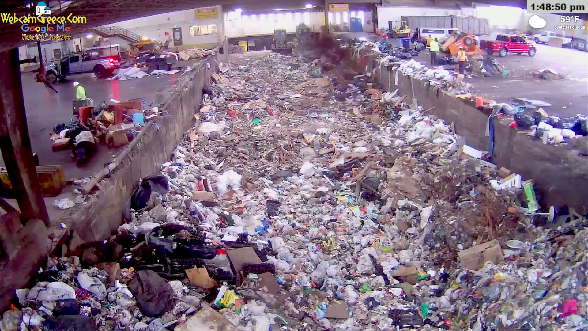 A large pile of trash sits in an outdoor waste facility under overcast skies, with several workers and vehicles visible nearby.