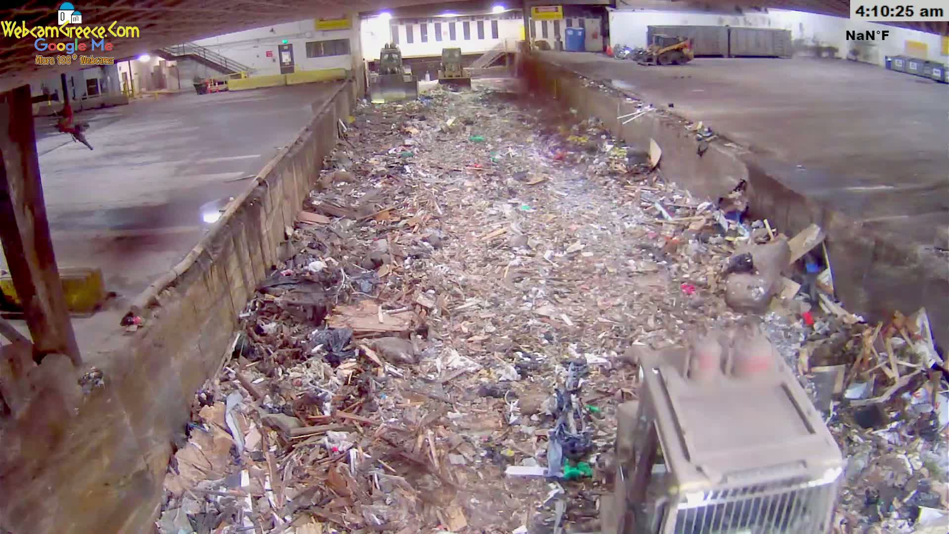 A large pile of trash and debris fills a long, narrow indoor space, with a piece of heavy machinery partially visible in the lower right corner.