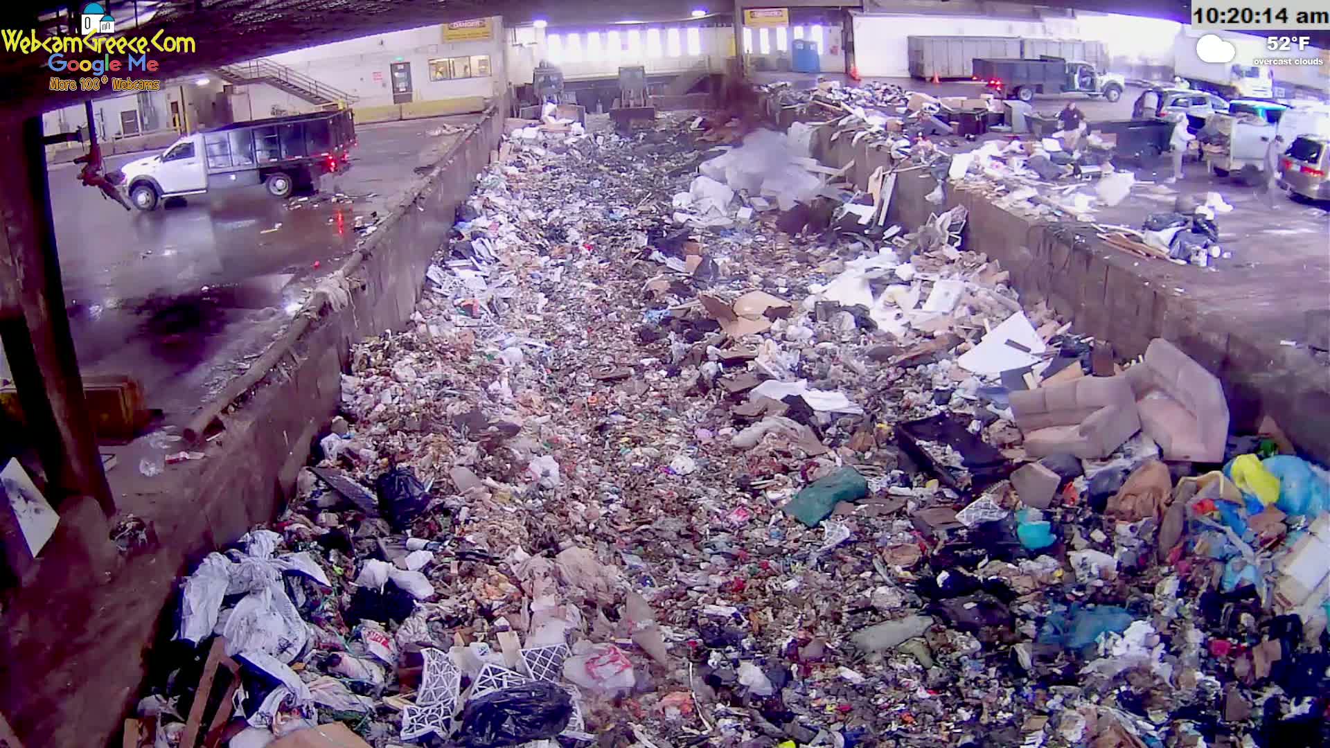 A large pile of refuse is seen under overcast skies in a waste disposal area, with a truck parked nearby and other vehicles visible in the background.