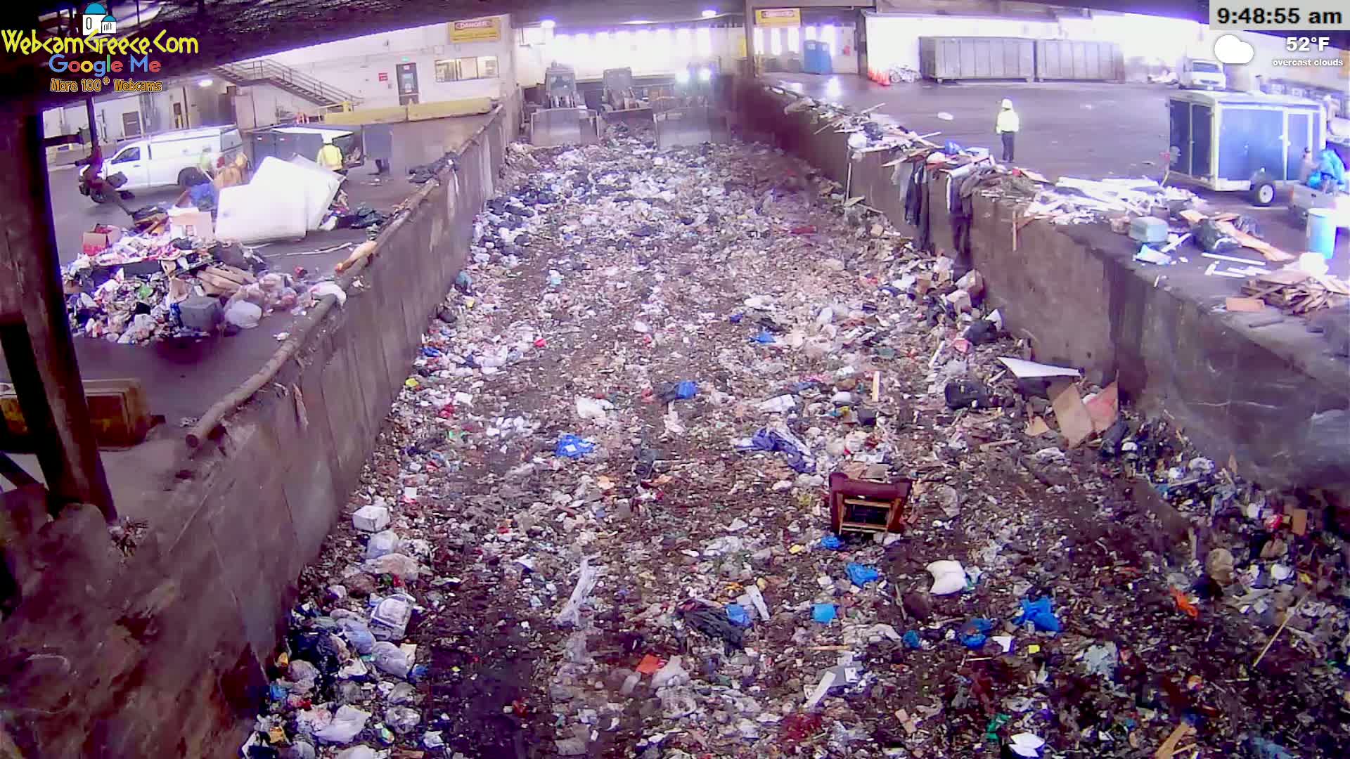 A large pile of trash fills a concrete-walled trench under overcast skies, with several people visible working nearby.