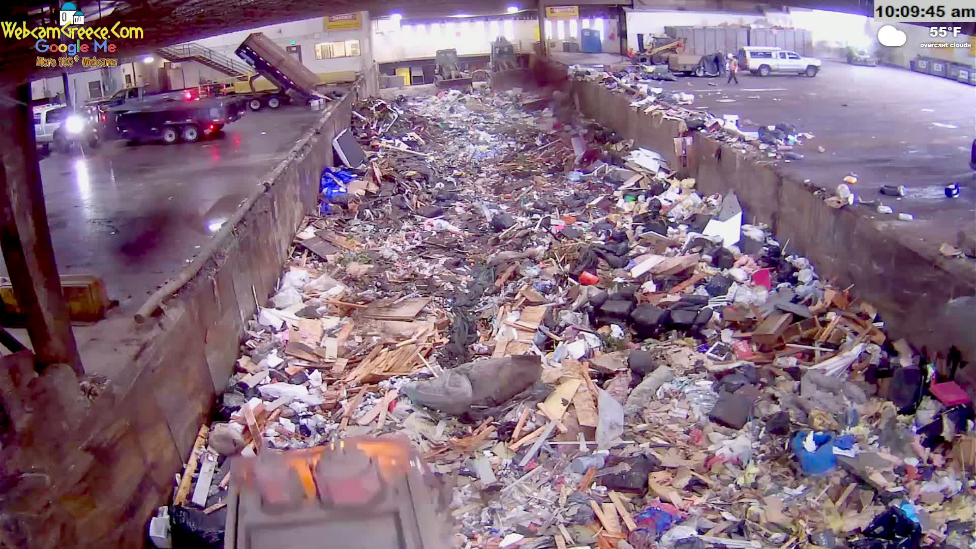 A large pile of refuse and debris fills a long, open-topped container under overcast skies.