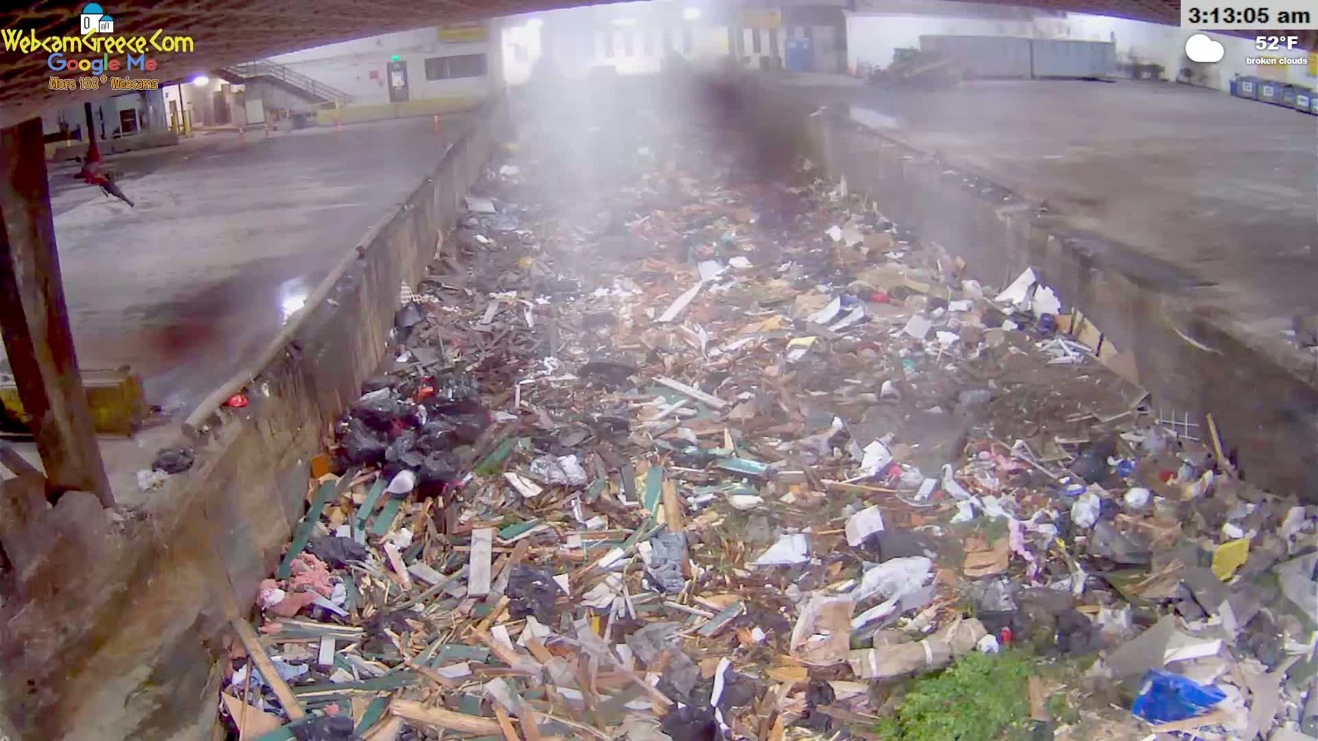 A large pile of debris, including wood and other refuse, fills a concrete trough-like area outdoors under broken clouds.