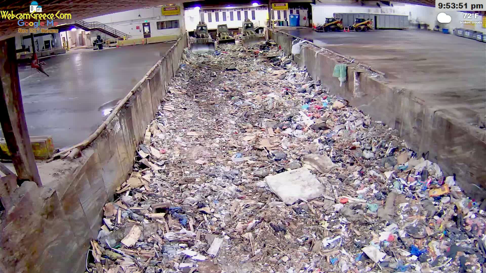 A large amount of refuse is piled inside a long, concrete trough, with two pieces of heavy machinery visible in the background under a broken cloud sky.