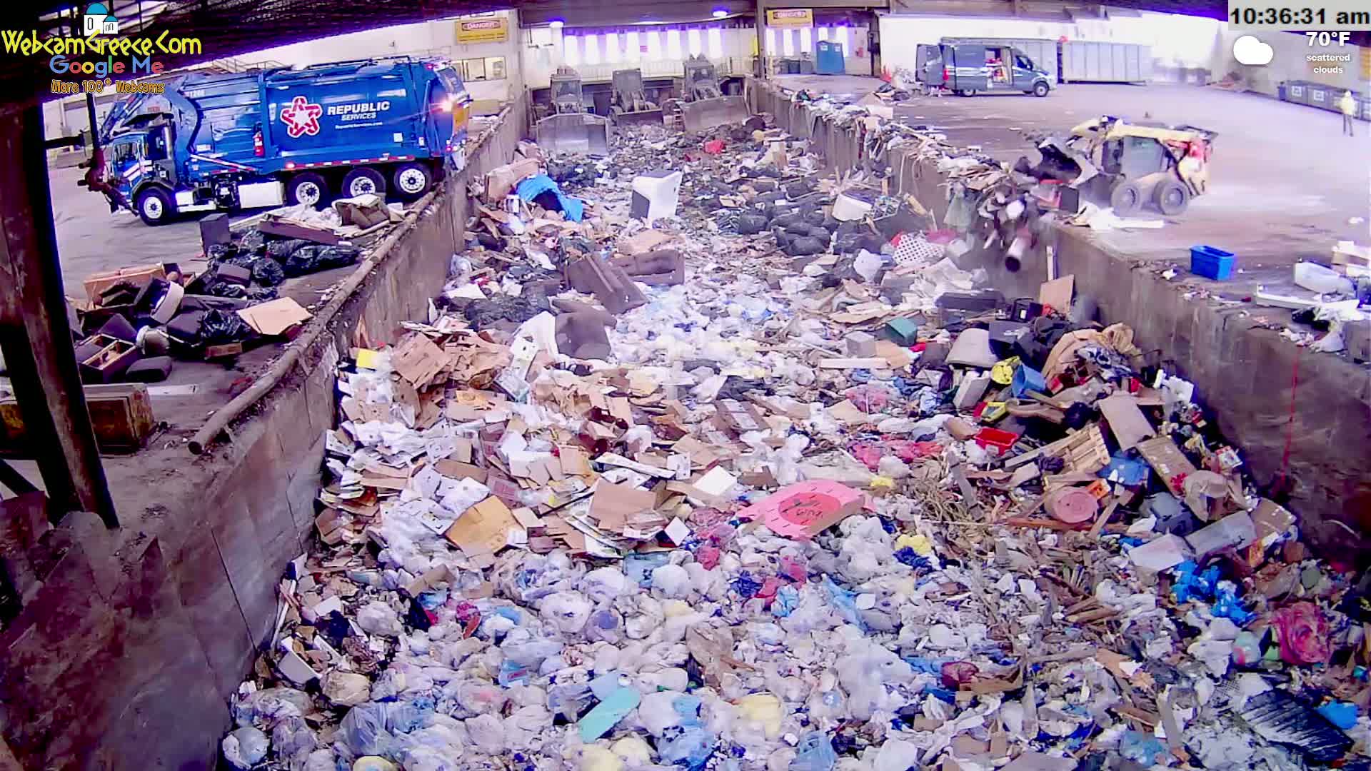 A large amount of trash and recyclables are piled high in a waste disposal area under a partly cloudy sky, with a garbage truck and a small bulldozer visible nearby.