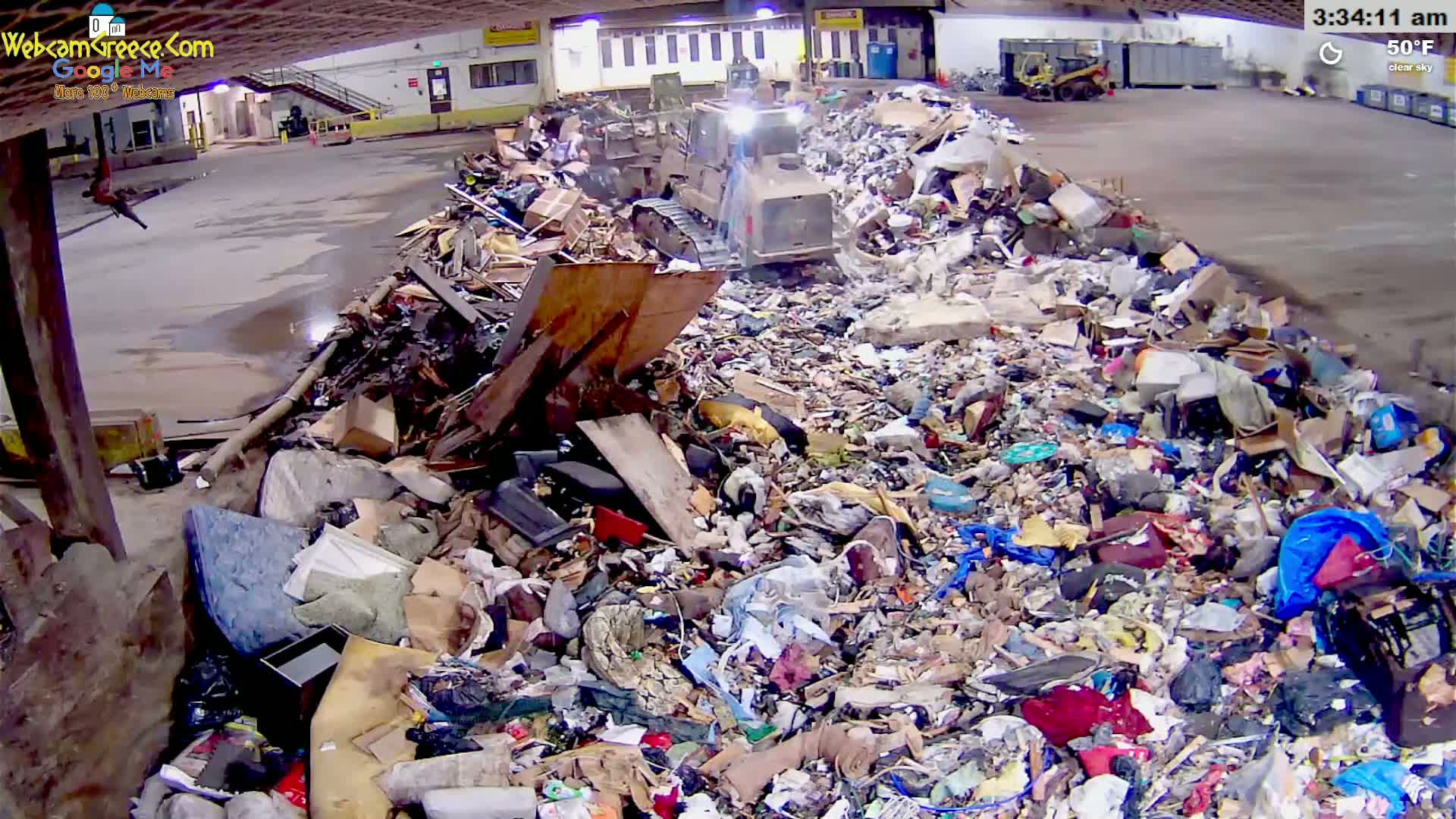 A bulldozer is compacting a large pile of trash and debris in an indoor facility on a clear, 50°F day.