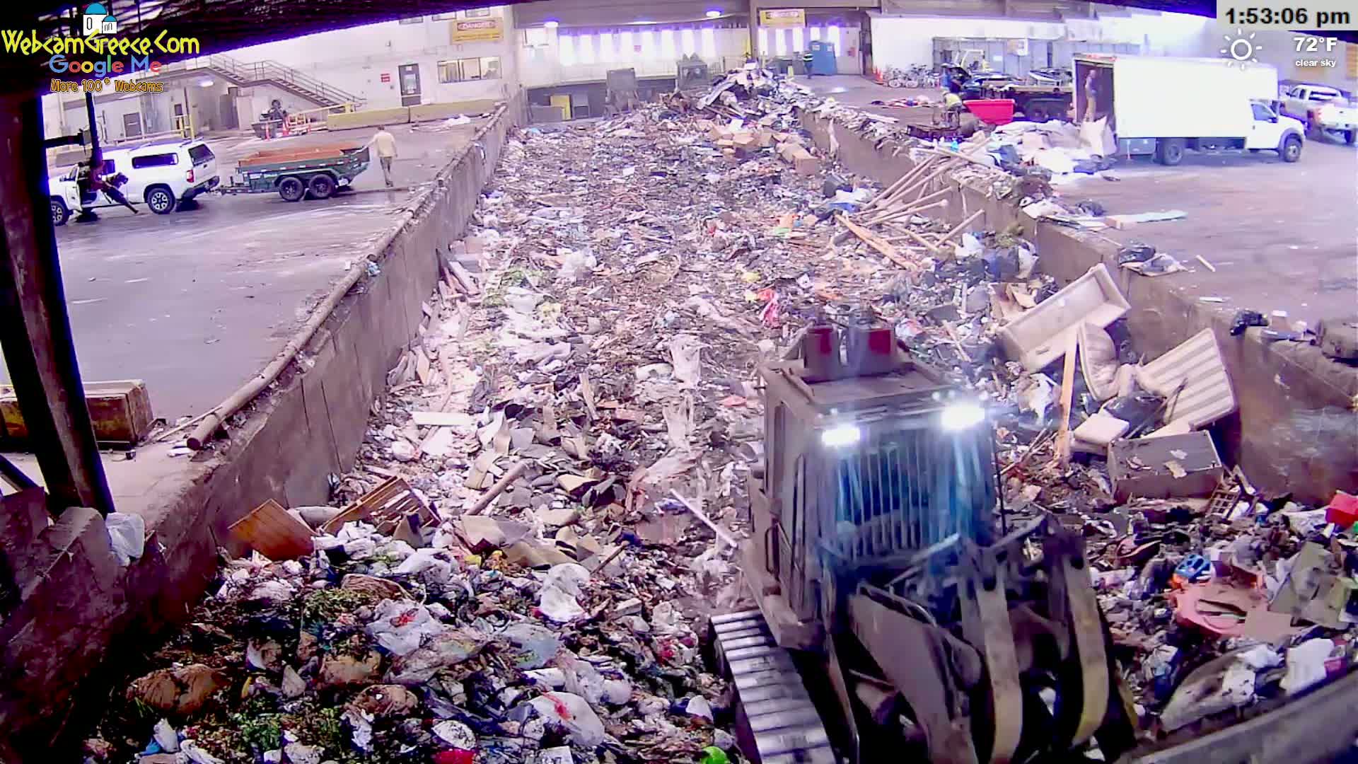 A bulldozer is compacting a large pile of trash and debris in an outdoor waste disposal area under a clear sky.