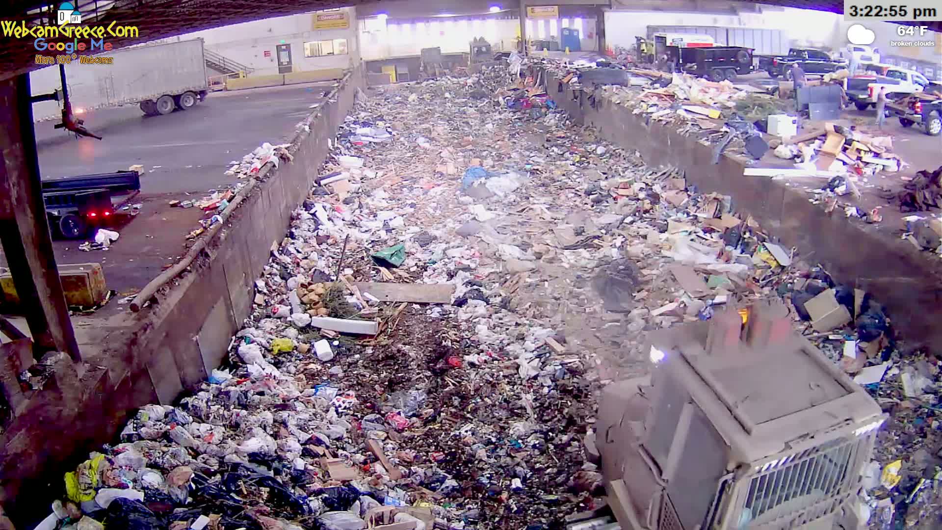 A large pile of garbage is being compacted by a machine in an outdoor area under broken clouds and a temperature of 64°F.