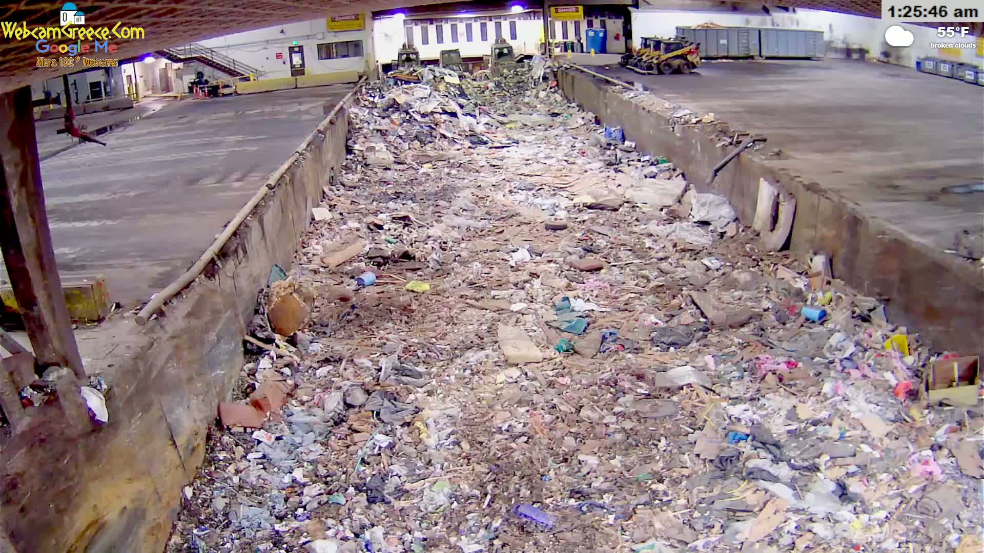 A large pile of trash and debris fills a long, open-topped container in an indoor facility under broken clouds at 55°F.