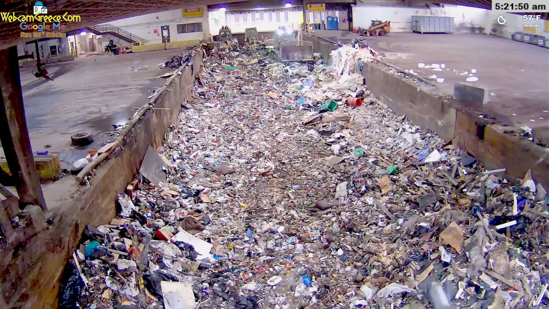 A large pile of mixed waste sits in an indoor waste processing facility under a clear sky.
