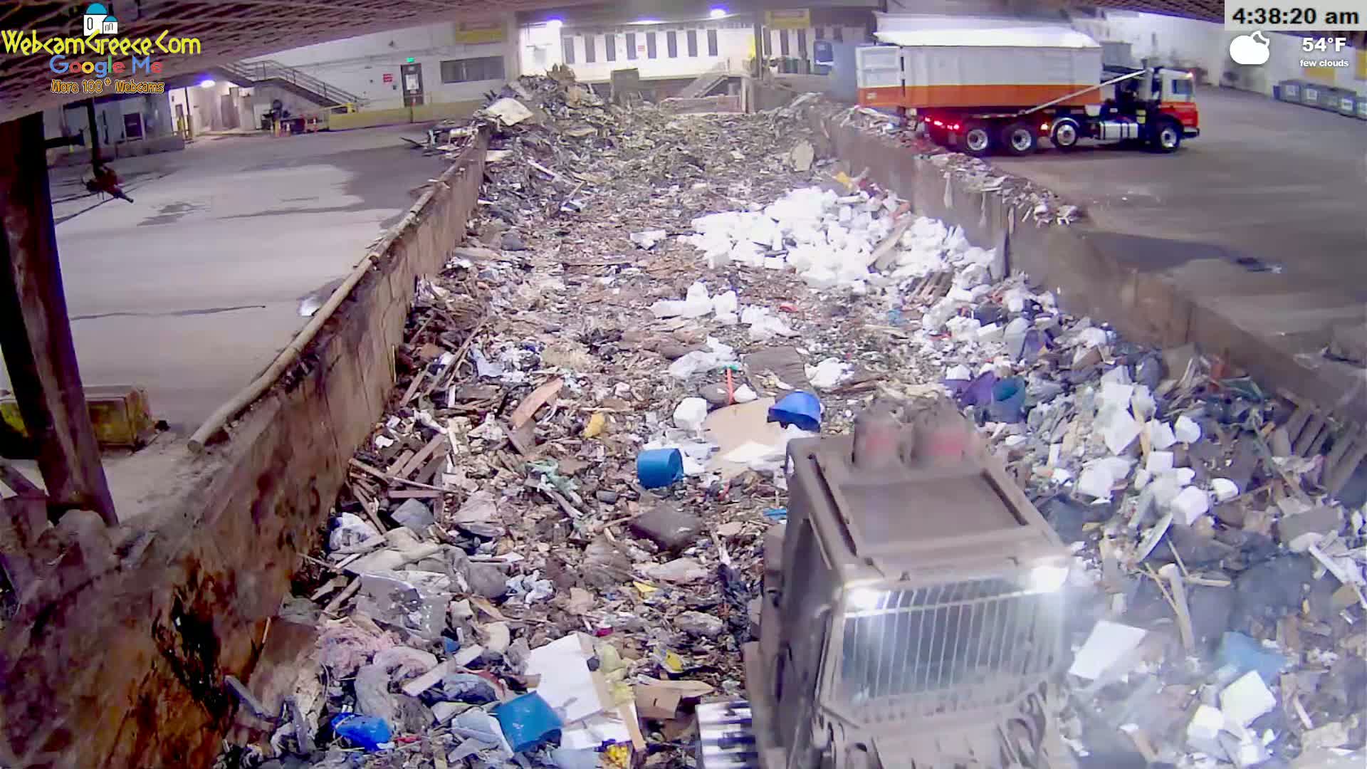 A large pile of garbage is seen inside a waste processing facility under few clouds at 54°F, with a bulldozer compacting the waste near a semi-truck.