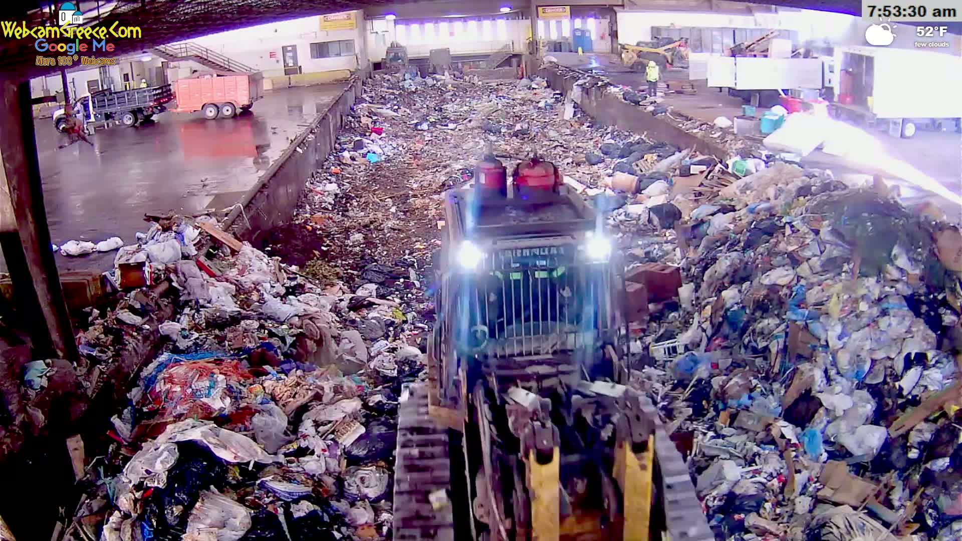 A bulldozer moves through a large pile of garbage in a waste processing facility under a partly cloudy sky.