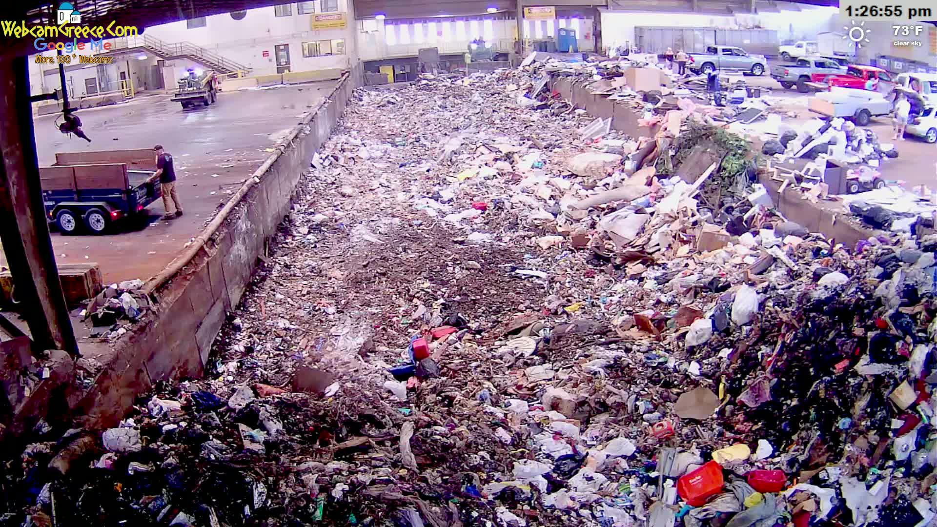 A large pile of trash is seen from an elevated view on a clear, 73°F day, with a person dumping more waste from a trailer into it.