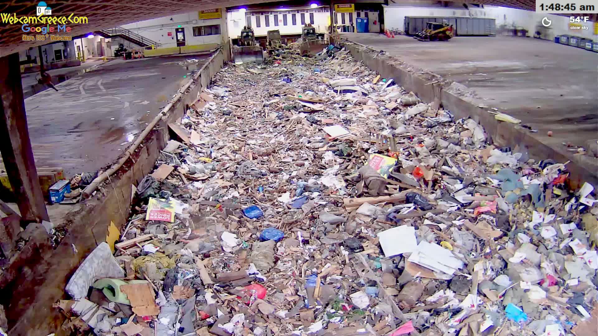 A large pile of mixed waste sits in an indoor waste facility under a clear sky at 54°F.