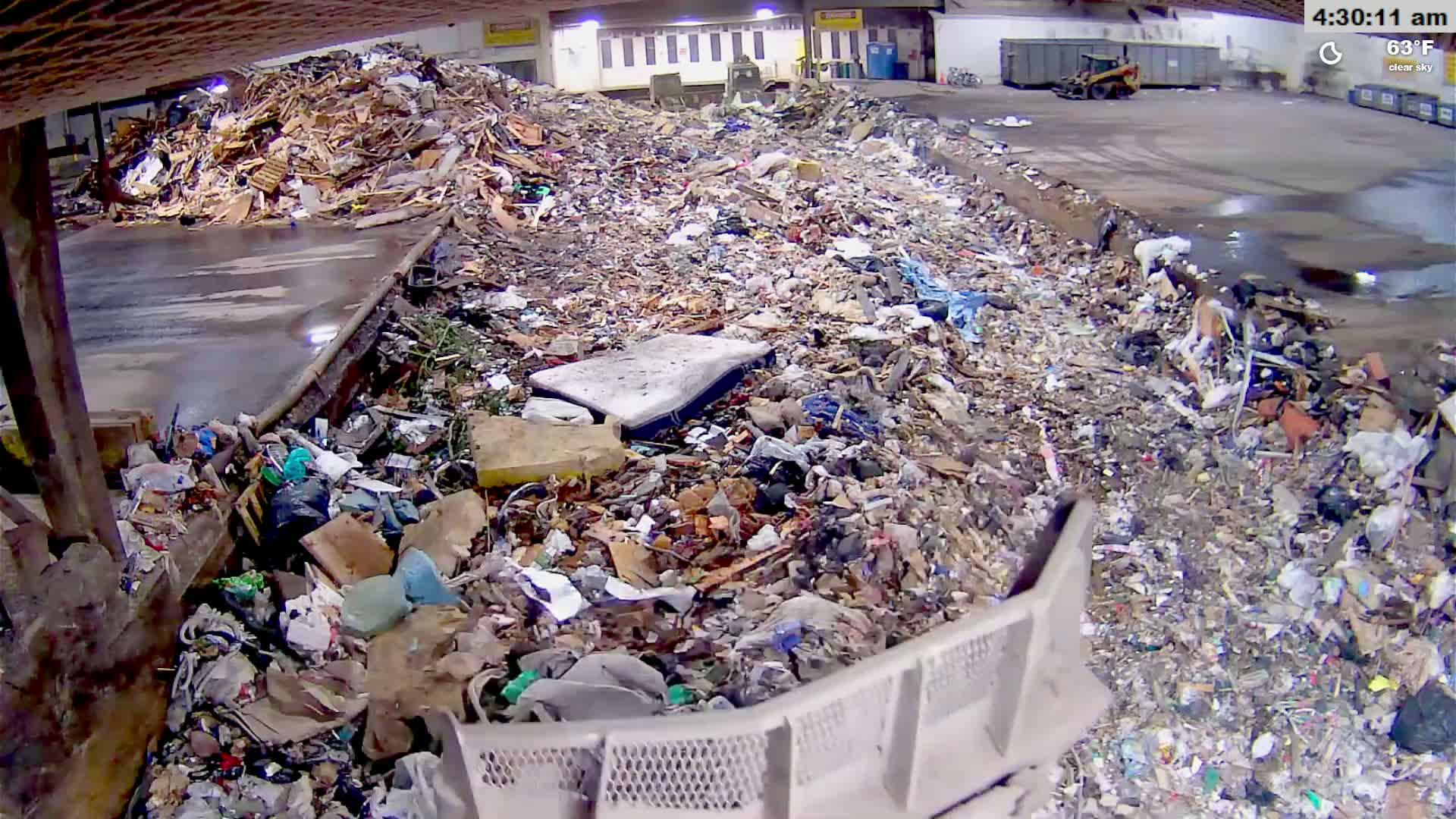 An indoor view of a large amount of garbage and debris piled high in a concrete trough, with two pieces of heavy machinery visible in the distance.