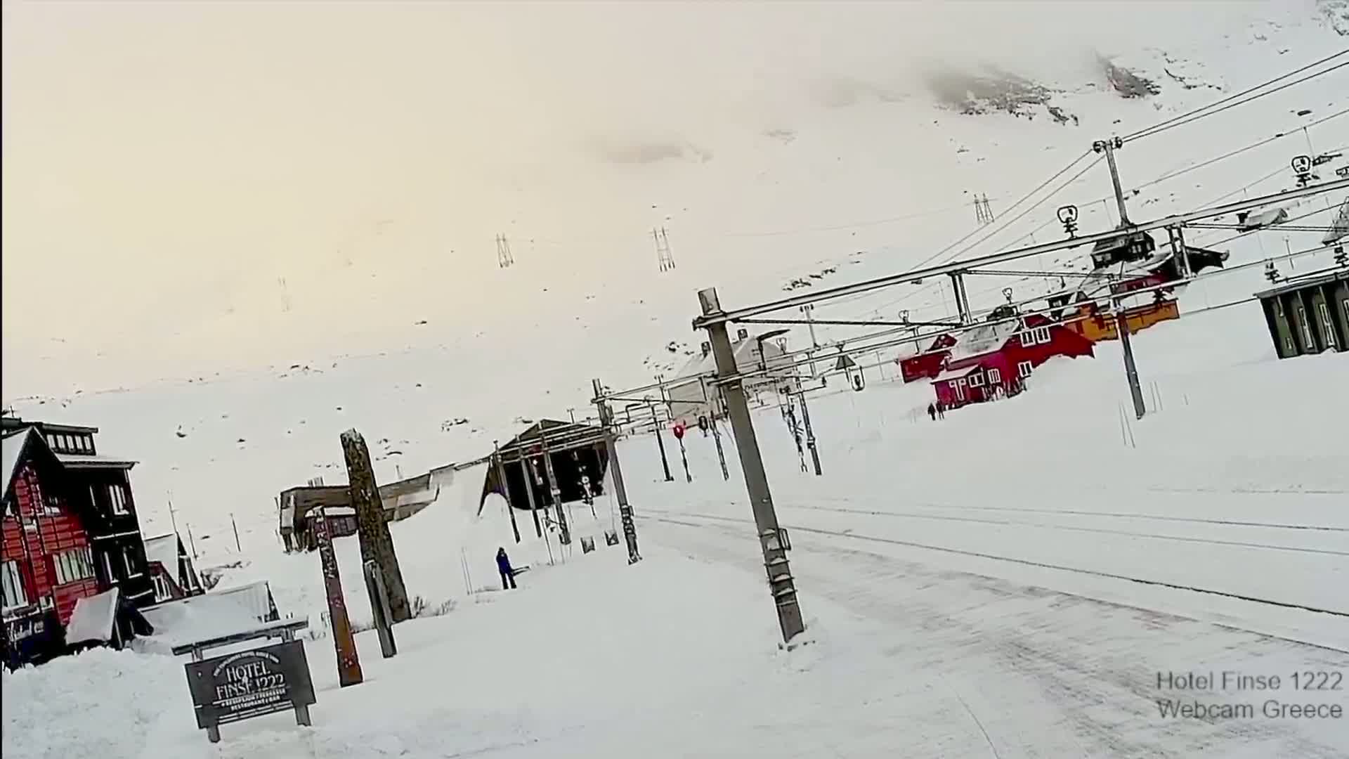 A snowy landscape shows a road and railway tracks next to a snow-covered mountain with several small buildings and a larger building, all under an overcast sky.