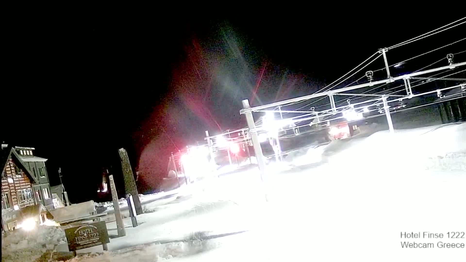 A nighttime snow scene shows a train passing illuminated overhead power lines near a snow-covered building.