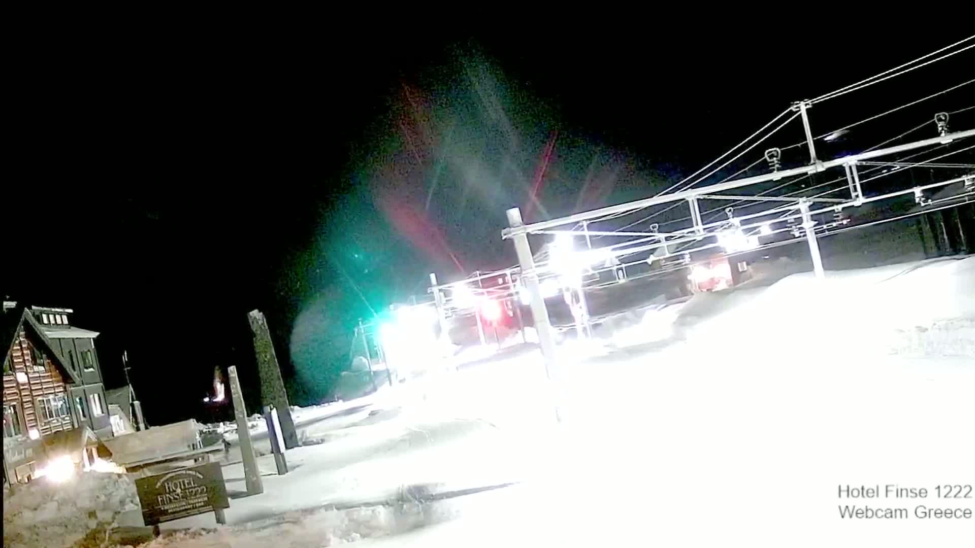 A nighttime snowy scene shows a train passing under overhead electrical lines near a wooden building, with bright lights illuminating the snow-covered ground.