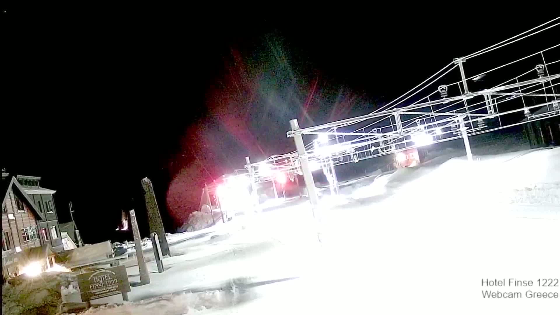 A ski lift illuminated at night under a dark sky sits in a snow-covered area near a building.