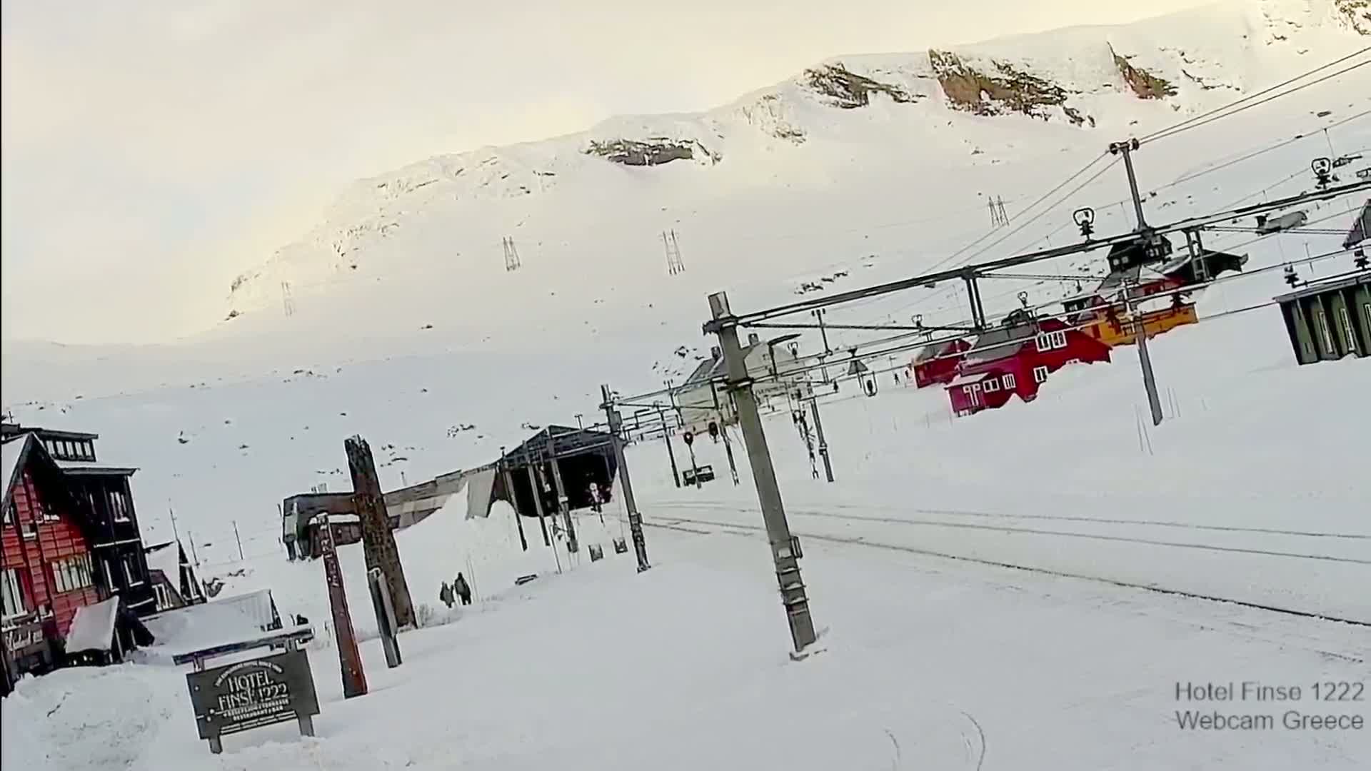 A snow-covered landscape with several red buildings near a ski lift,  a mountain in the background under a mostly cloudy sky.