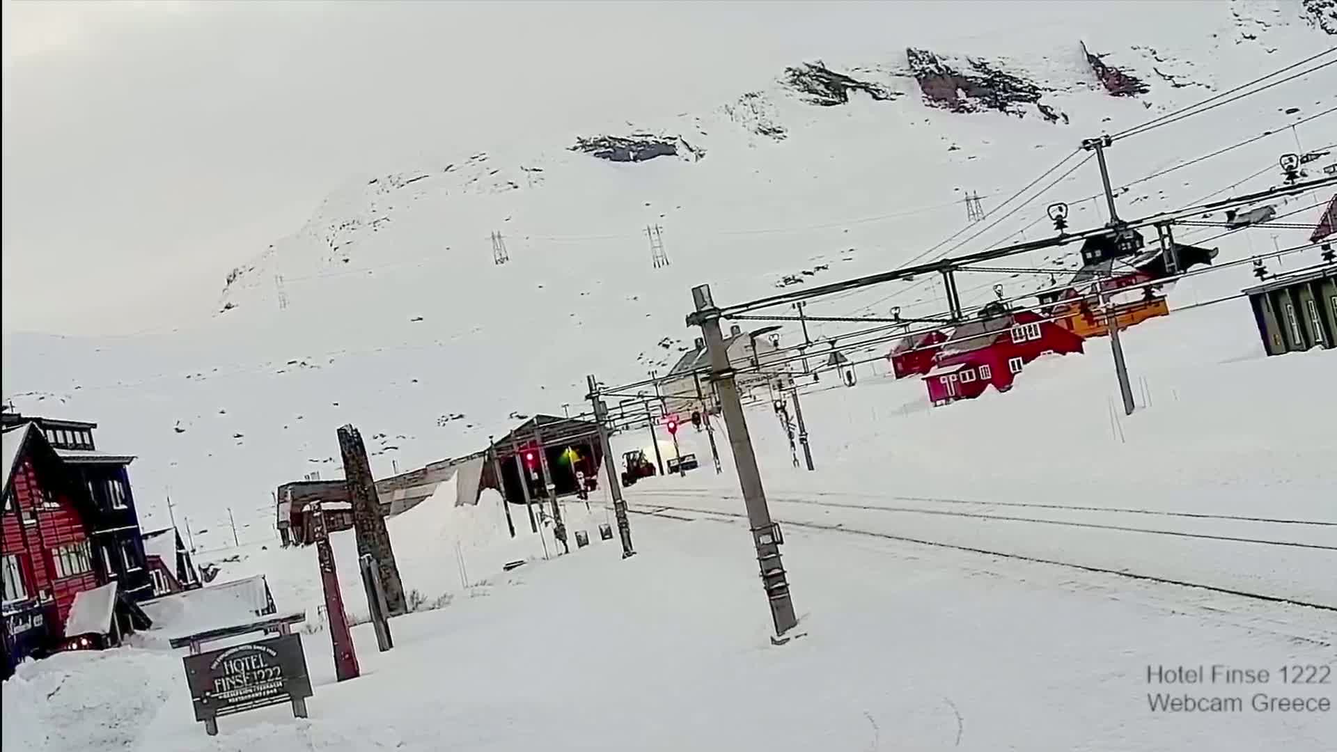A train approaches a snow-covered train station nestled in a mountainous region during a heavy snowfall.