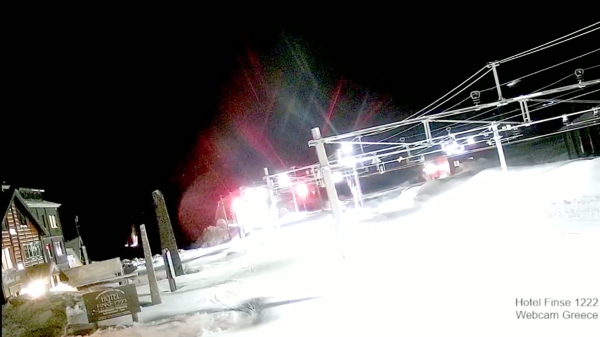 A nighttime view of a snow-covered landscape with overhead wires, a building with lights on, and red and white lights in the distance.