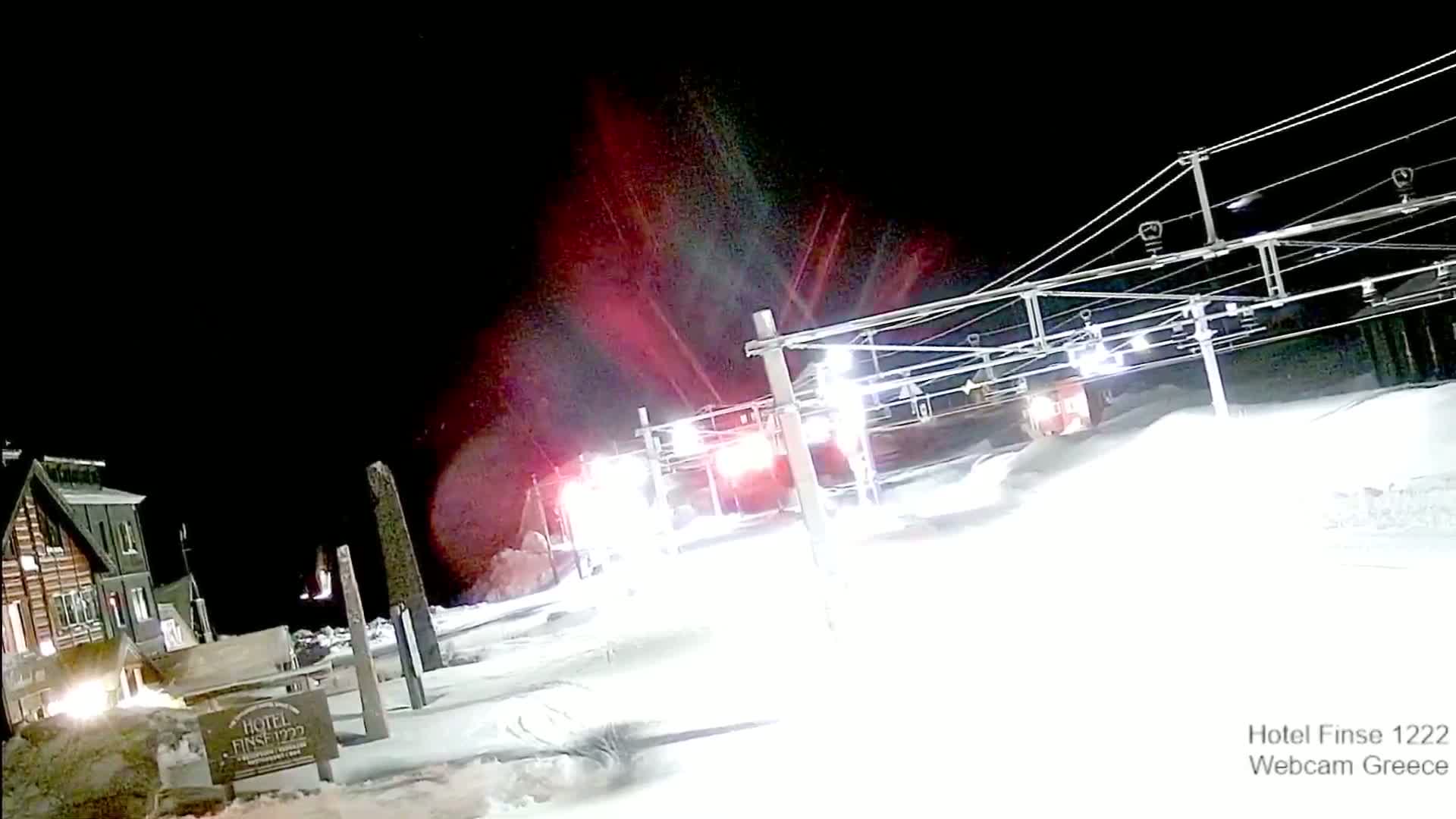 At night, under a dark sky, a snow-covered ski lift and a building are illuminated, with red lights visible in the distance.