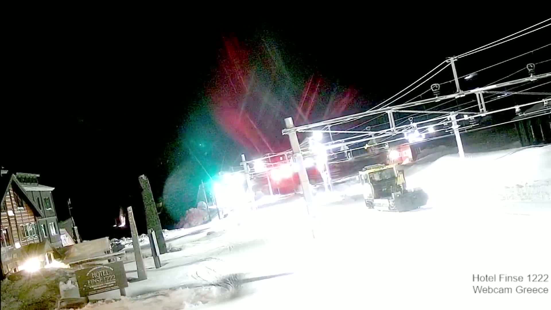 At night, a snowcat is grooming a snow-covered ski slope under bright lights, with a building and ski lift infrastructure visible in the background.