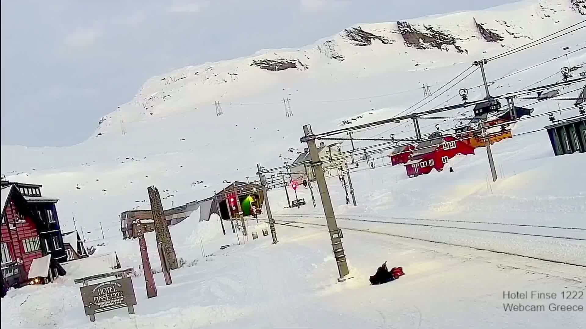 A snow-covered landscape with a train track, several buildings, and a person sitting on what appears to be a snowmobile near the tracks under a bright, mostly clear sky.