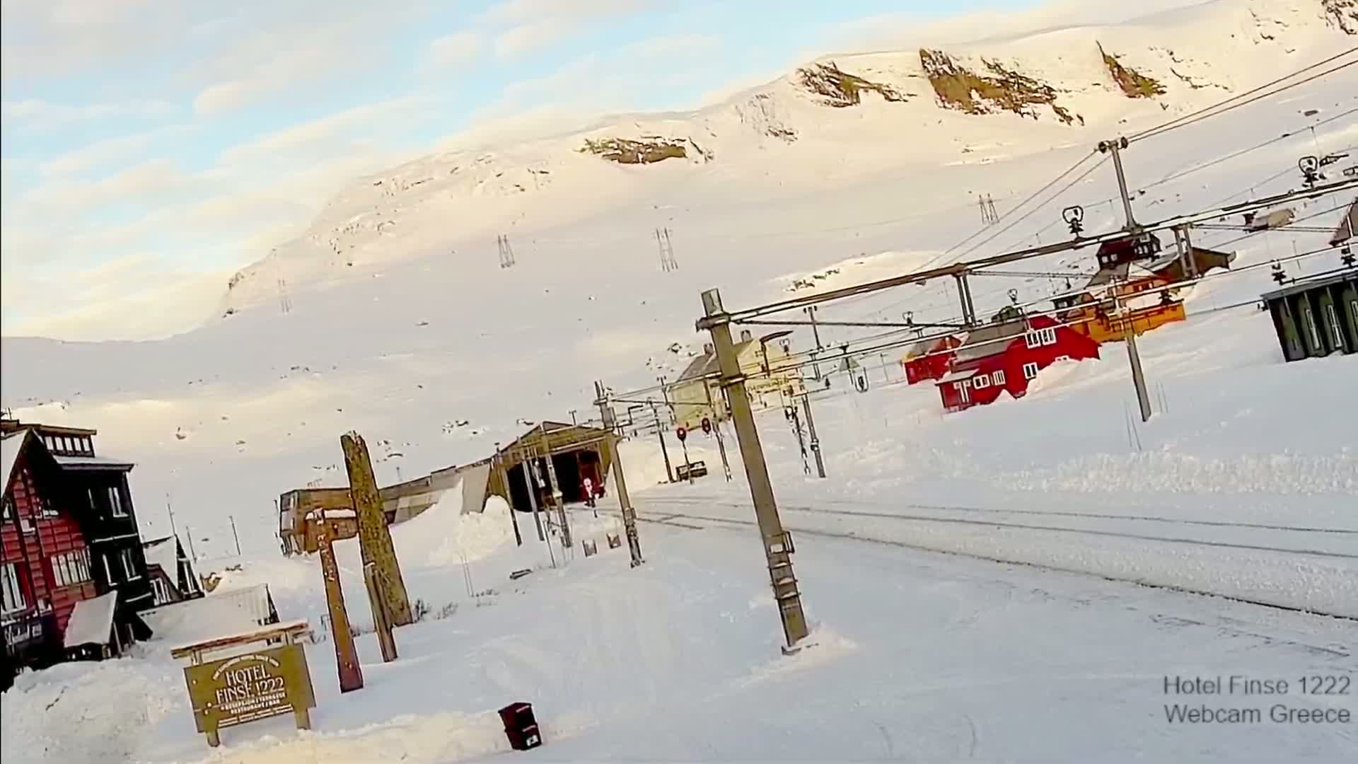 A snow-covered train station with red buildings and a mountain in the background under a partly cloudy sky.