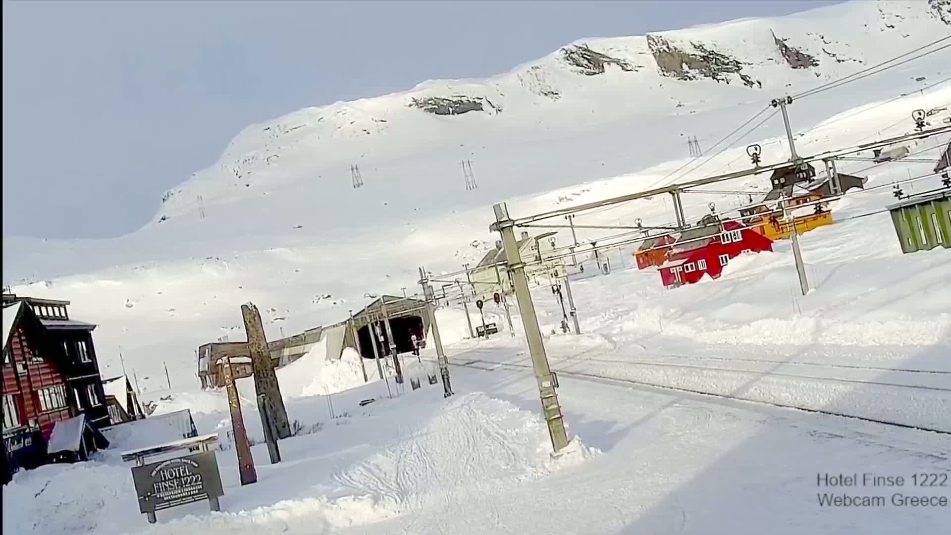 A snow-covered mountainside scene features a train track winding past snow-covered buildings and a tunnel entrance, under a bright, clear sky.