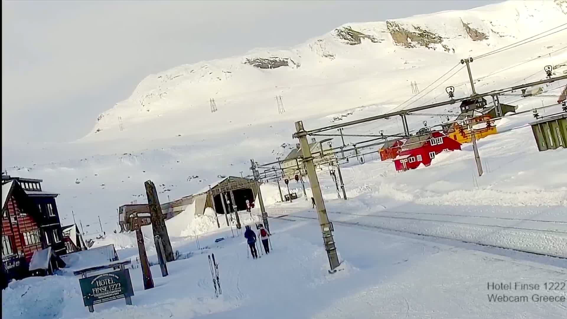 Two cross-country skiers walk along a snowy path near a mountain, past buildings and ski lift infrastructure under a bright, clear sky.