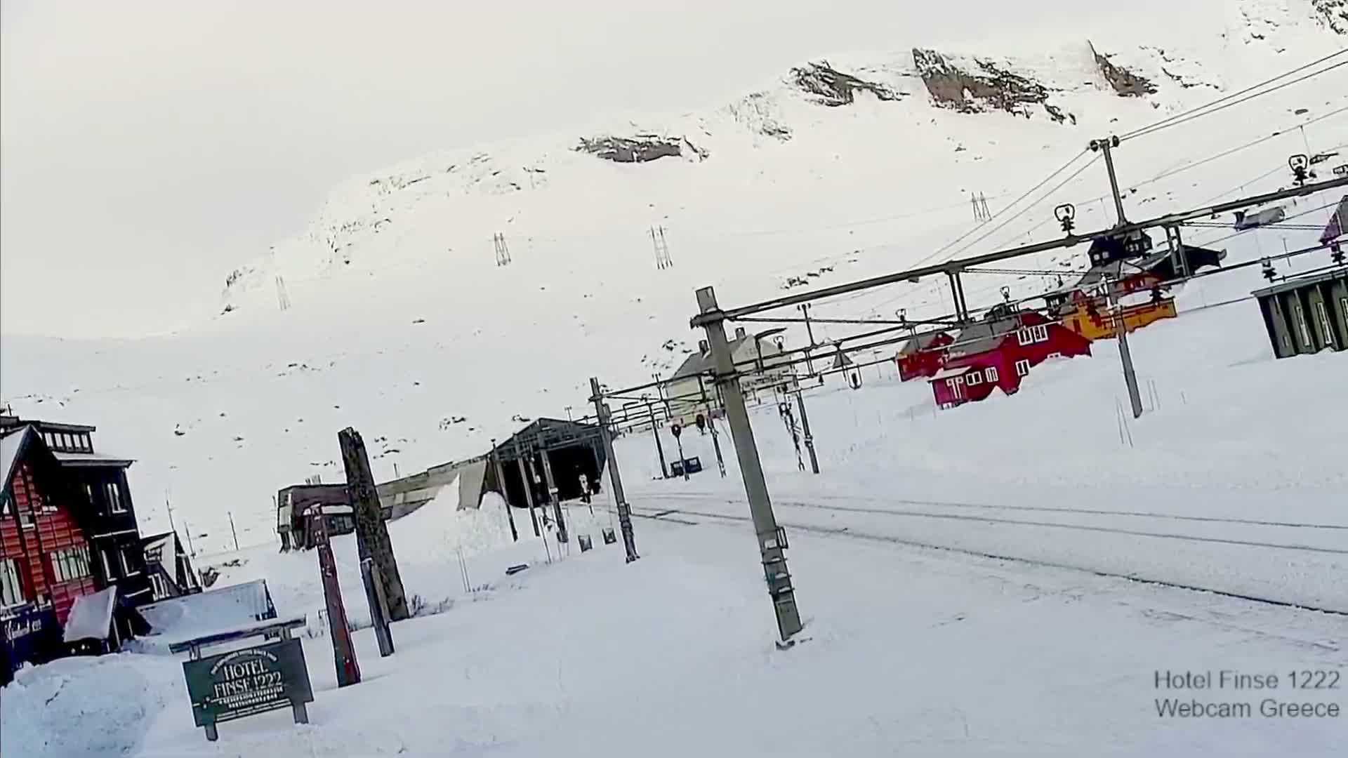 A snowy mountainside scene shows a road and several small buildings partially buried in snow, with ski lift infrastructure and a tunnel entrance visible.