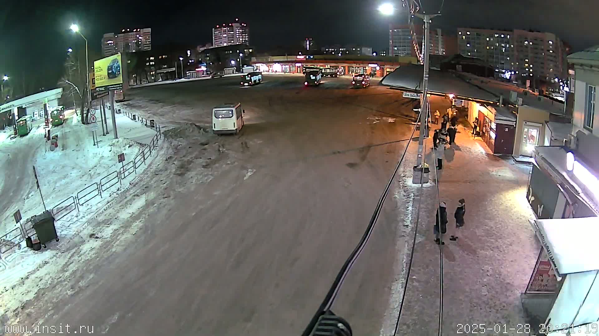 A nighttime, snow-covered bus stop with several buses and people standing around, under streetlights.