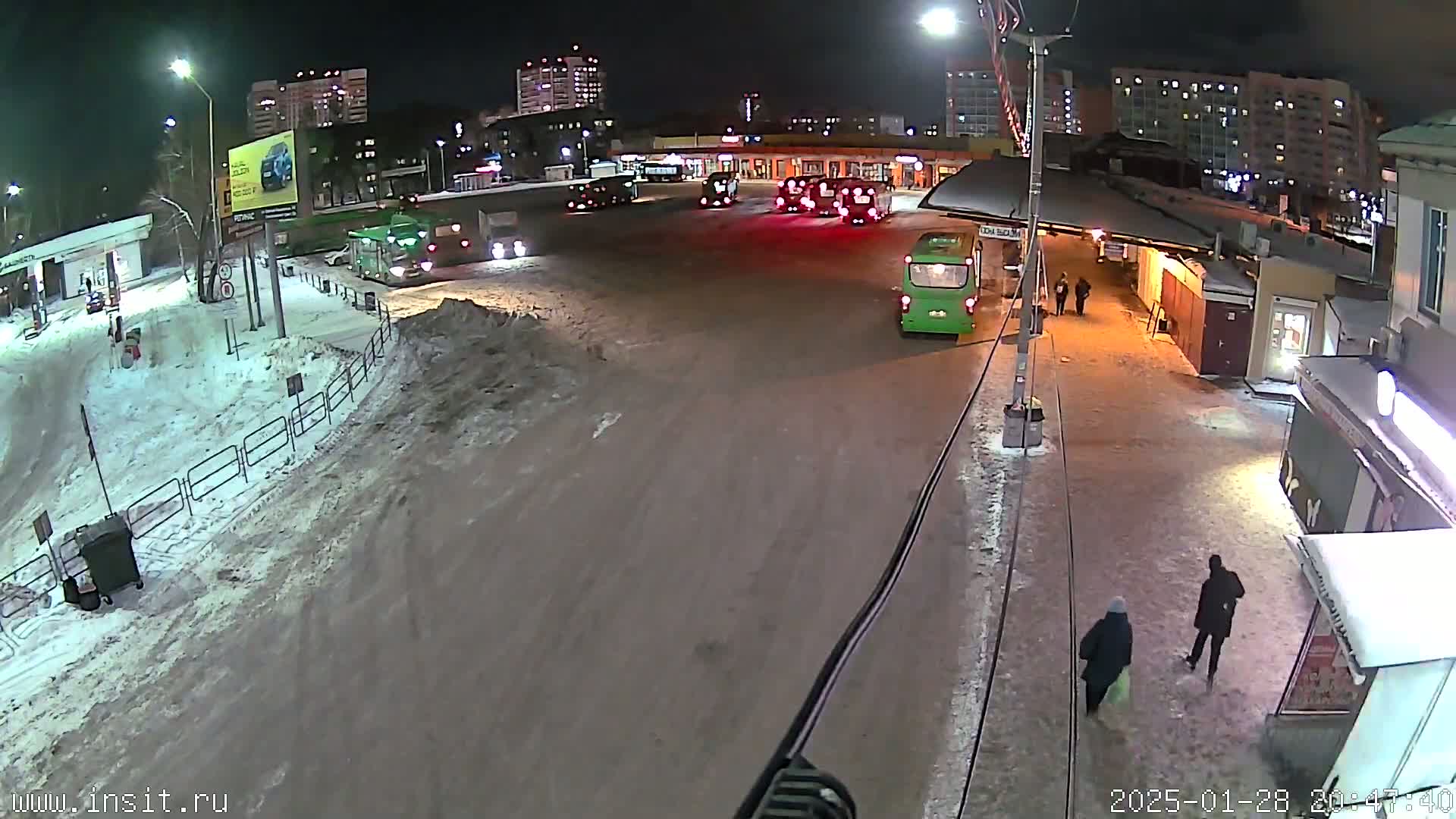 A nighttime, snowy scene shows several buses parked at a terminal, with a few pedestrians walking nearby.