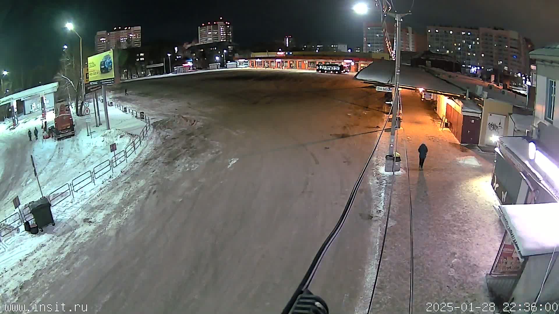 A nighttime, snowy scene shows a mostly empty paved area with a few buildings, a bus, and a single pedestrian.
