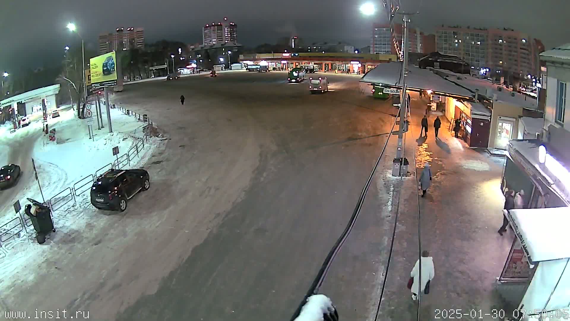 A snow-covered nighttime scene shows a bus stop with several people walking near buildings and parked cars.