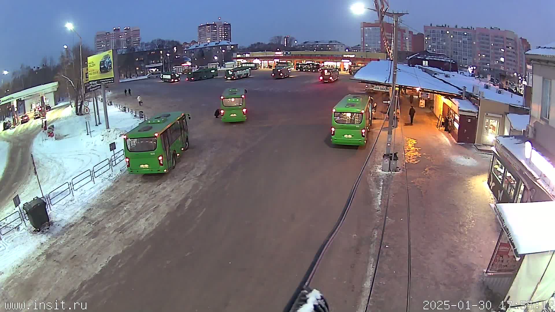 Several green buses are parked in a snowy bus depot at dusk, with surrounding buildings visible in the background.