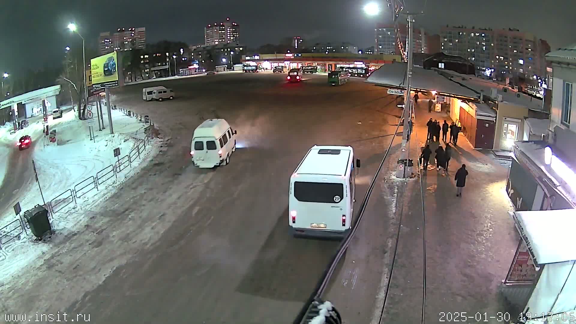 A nighttime scene of a snowy bus stop with several vehicles and people present.