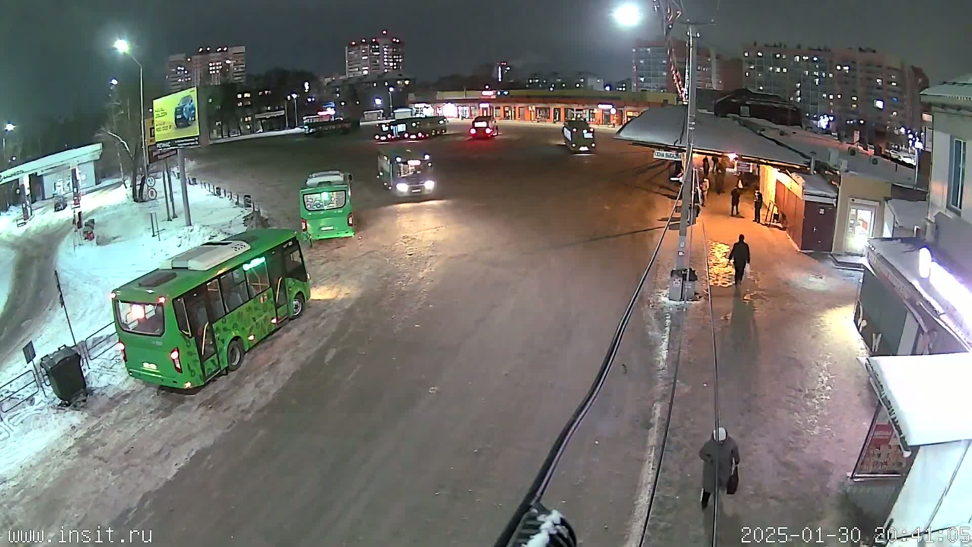 Several buses and a few people are at a snowy bus stop at night.